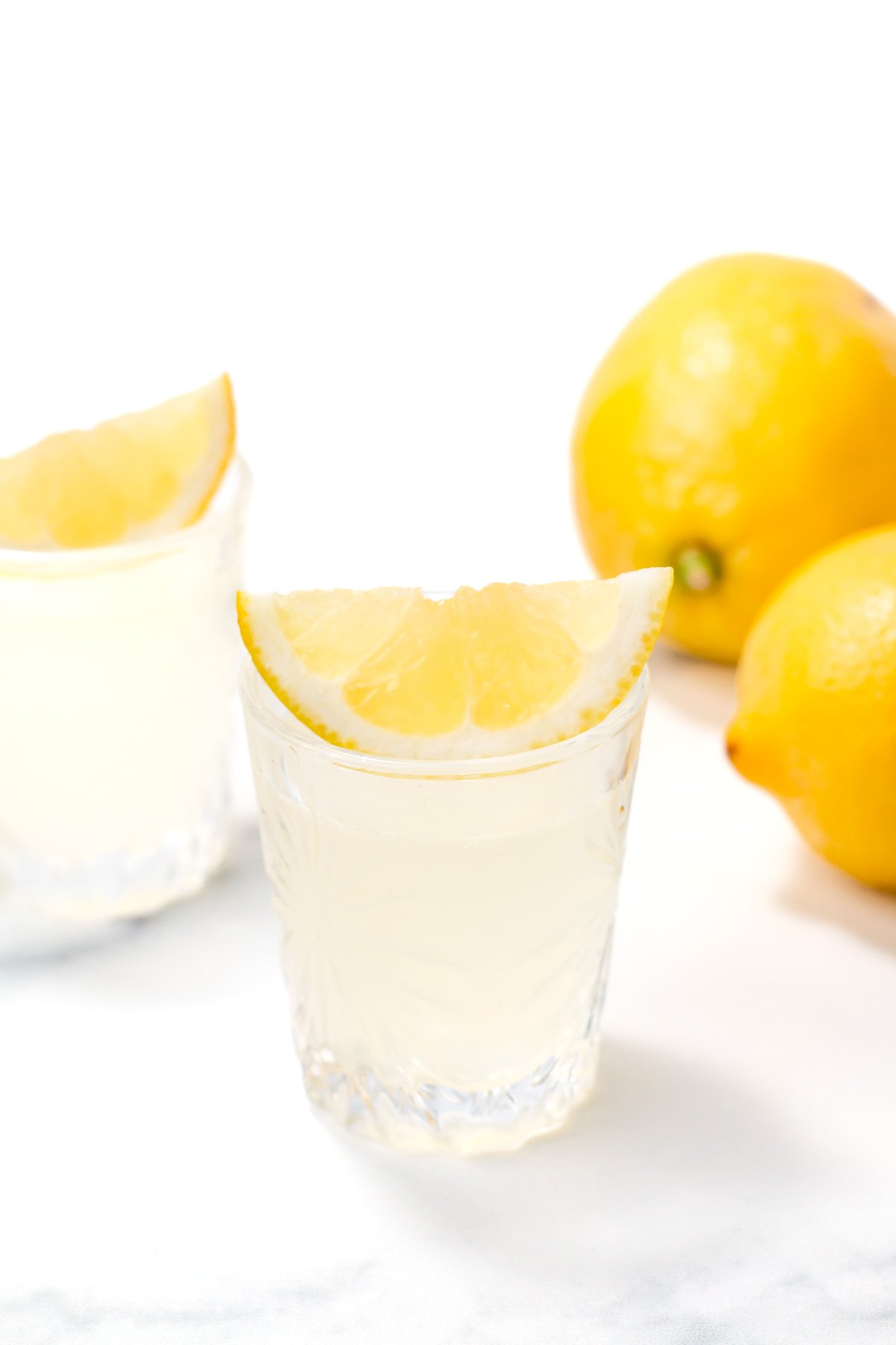 A glass of lemon juice with a lemon wedge on the rim, another glass, and whole lemons in the background on a white surface.