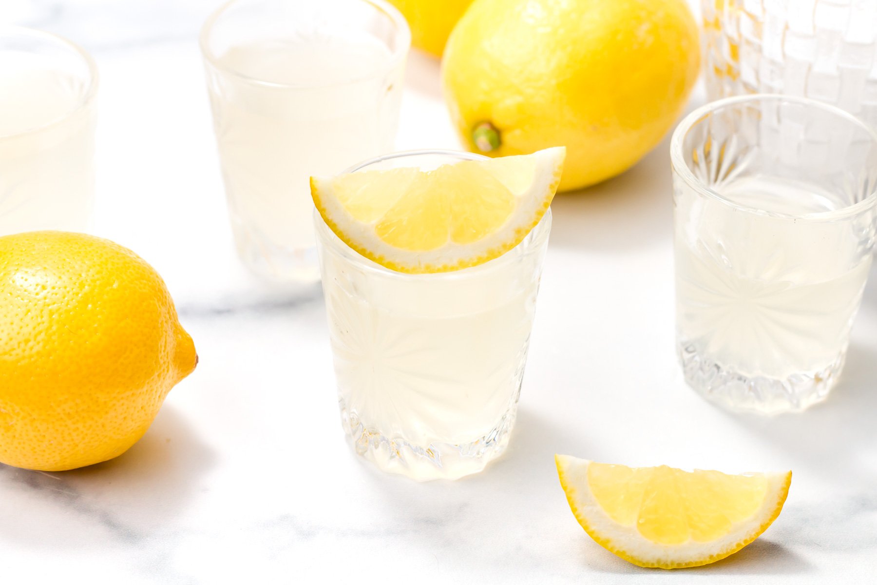 Several glasses of lemonade with lemon wedges, surrounded by whole and sliced lemons on a white surface.