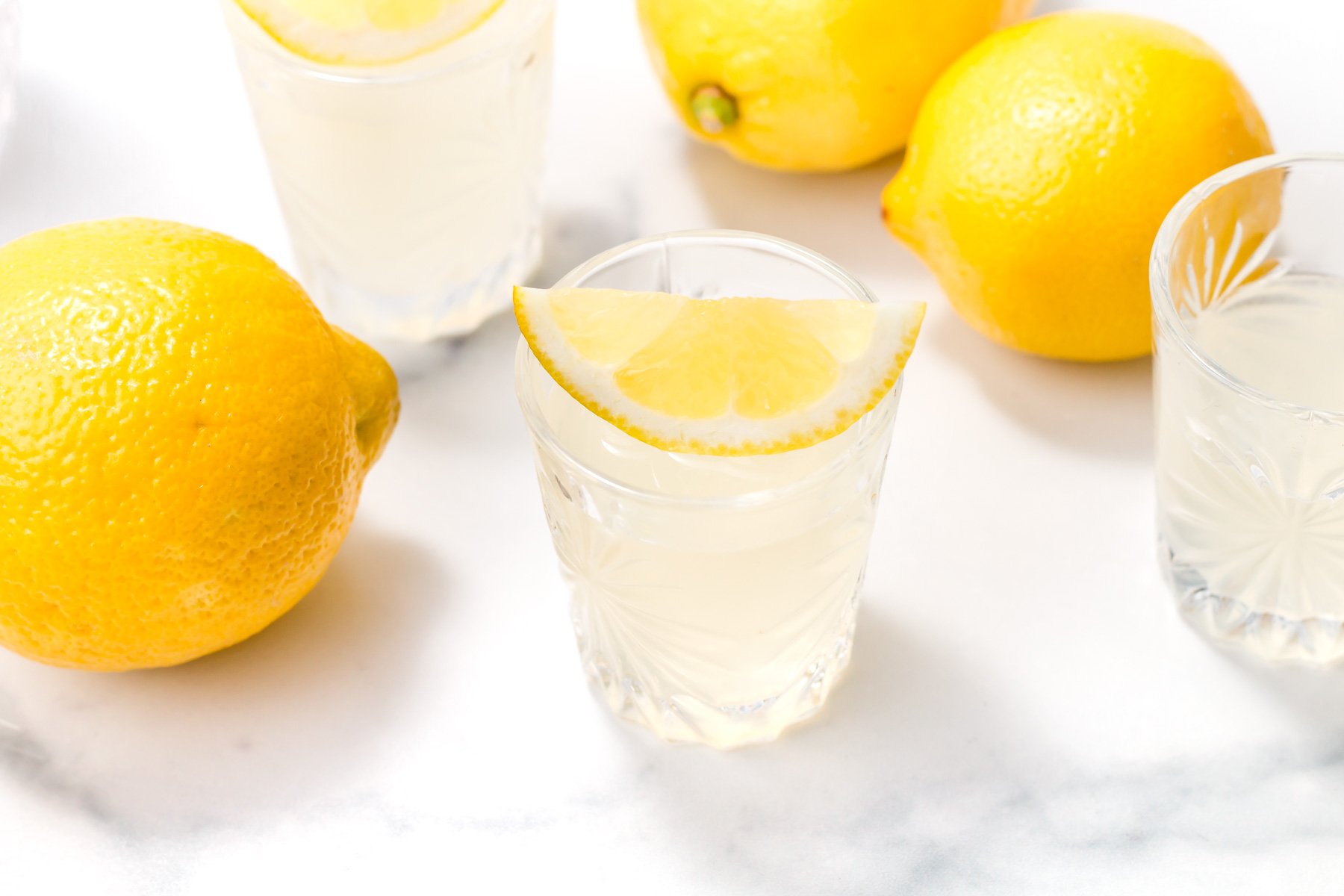 Three clear glasses of lemonade, one garnished with a lemon slice, surrounded by whole lemons on a white surface.