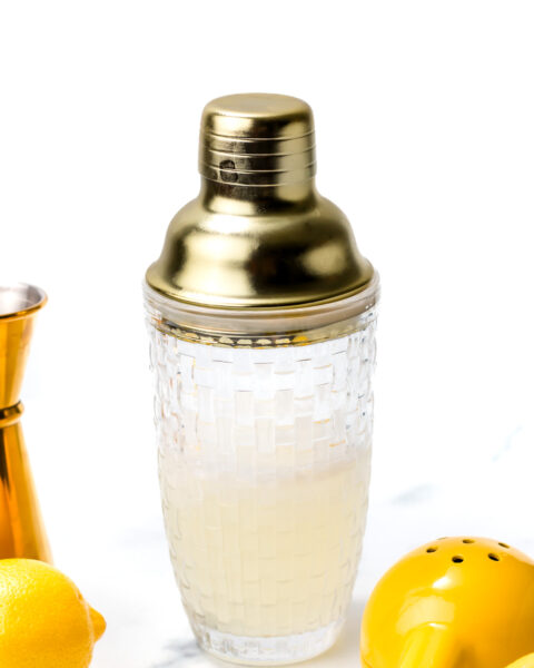 A glass cocktail shaker with a gold lid filled with a pale liquid, surrounded by a jigger, lemons, and a yellow citrus squeezer on a white background.