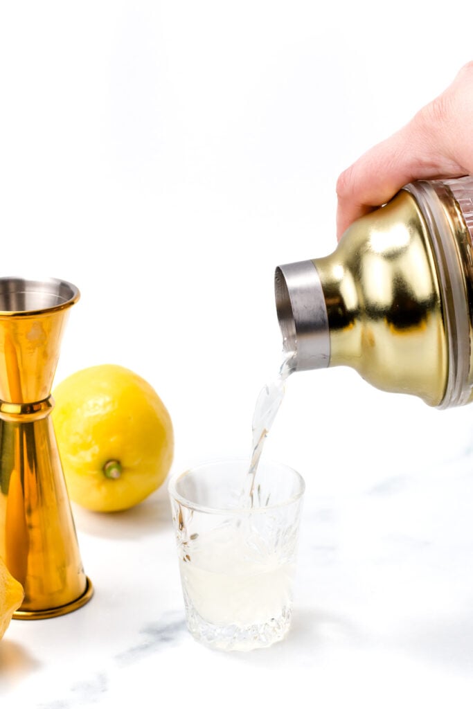 A hand pours liquid from a gold cocktail shaker into a glass, with a jigger and lemons nearby on a white surface.