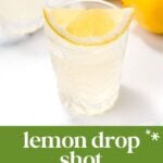 A clear glass of lemon drop shot with a lemon wedge on the rim, surrounded by fresh lemons. The zingy, bright, and smooth drink gets a colorful twist from hibiscus syrup.