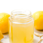 A hexagonal glass jar filled with light yellow liquid, surrounded by whole lemons and a gold spoon on a white surface.