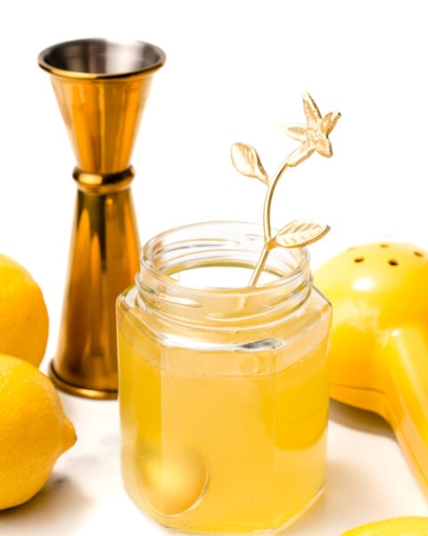 A glass jar filled with a yellow drink and a gold floral stirrer, surrounded by lemons, a yellow citrus squeezer, and a gold jigger on a white background.