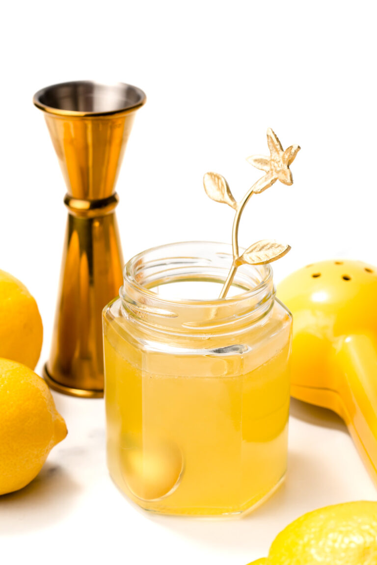 A glass jar filled with a yellow drink and a gold floral stirrer, surrounded by lemons, a yellow citrus squeezer, and a gold jigger on a white background.