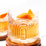 A glass of whiskey with ice and an orange peel garnish sits on stacked wooden coasters against a white background.