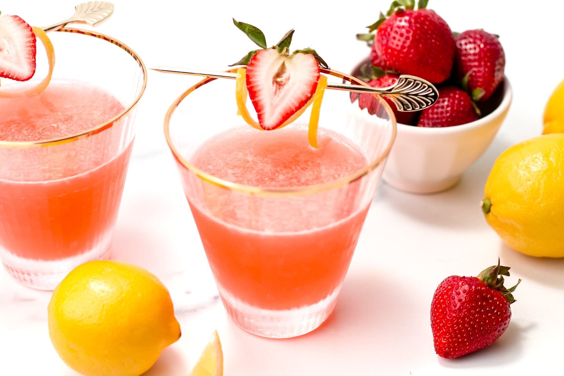 Two glasses of pink strawberry lemonade with lemon and strawberry garnish, surrounded by fresh strawberries and lemons on a white surface.
