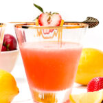 A glass of pink citrus drink garnished with a strawberry and lemon slice, surrounded by fresh strawberries and lemons on a white surface.