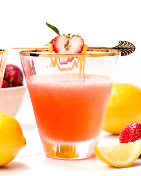 A glass of pink citrus drink garnished with a strawberry and lemon slice, surrounded by fresh strawberries and lemons on a white surface.