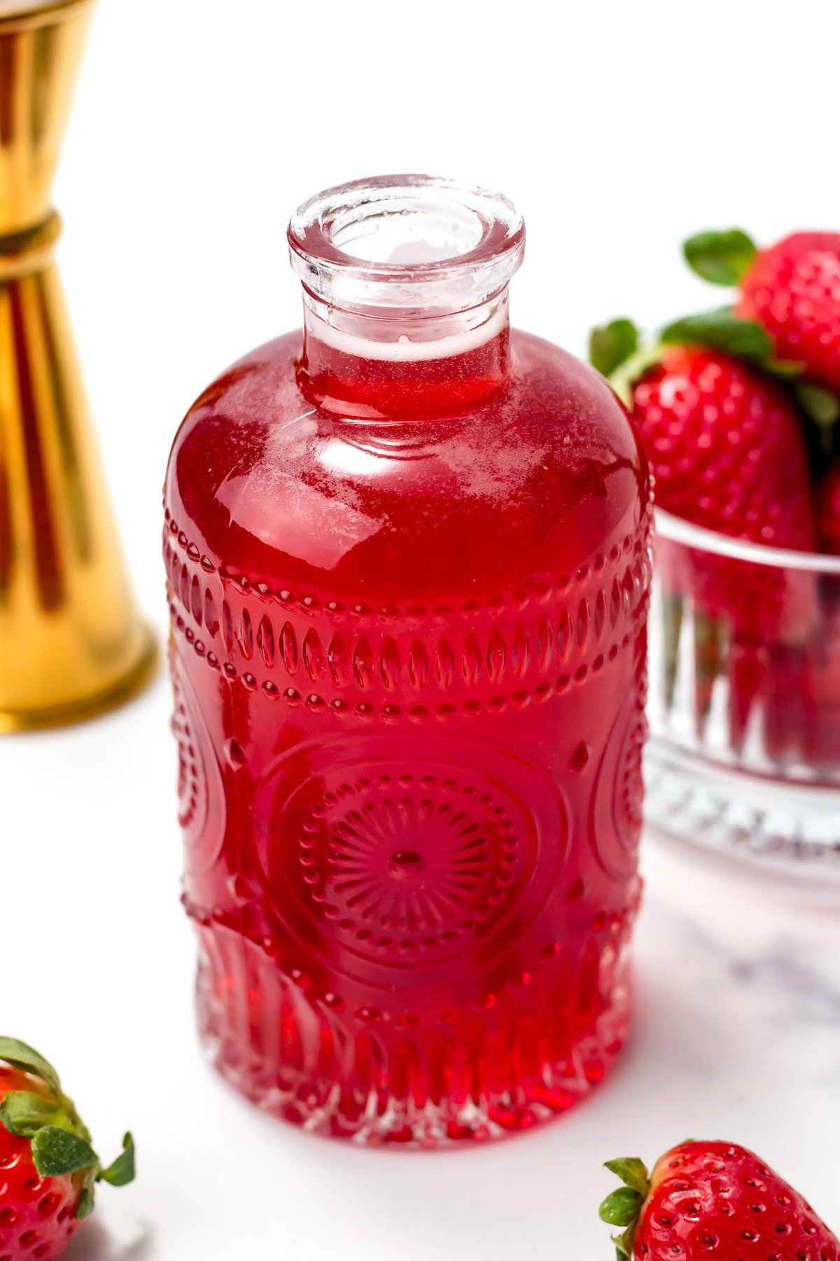 A decorative glass bottle filled with red liquid sits next to fresh strawberries and a glass container, all on a white surface.