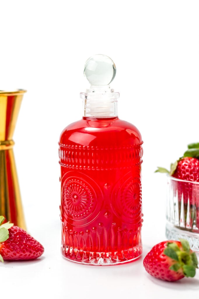A decorative glass bottle filled with red liquid, surrounded by fresh strawberries and a metal jigger, on a white background.
