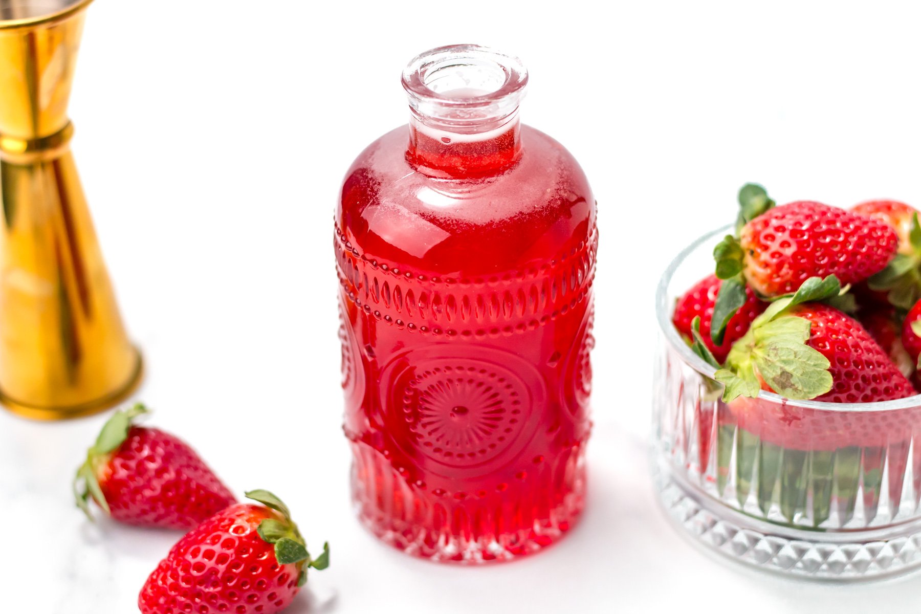 A decorative glass bottle filled with red liquid sits beside a glass bowl and loose strawberries on a white surface.