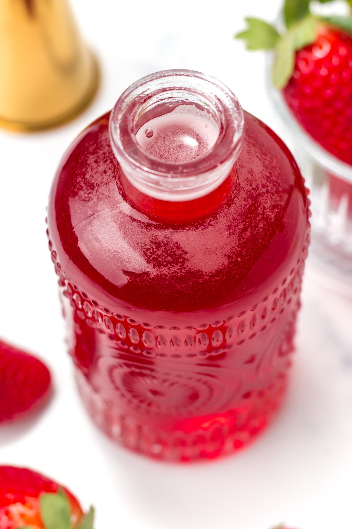 A clear glass bottle filled with red liquid, placed on a white surface with strawberries and a gold jigger nearby.