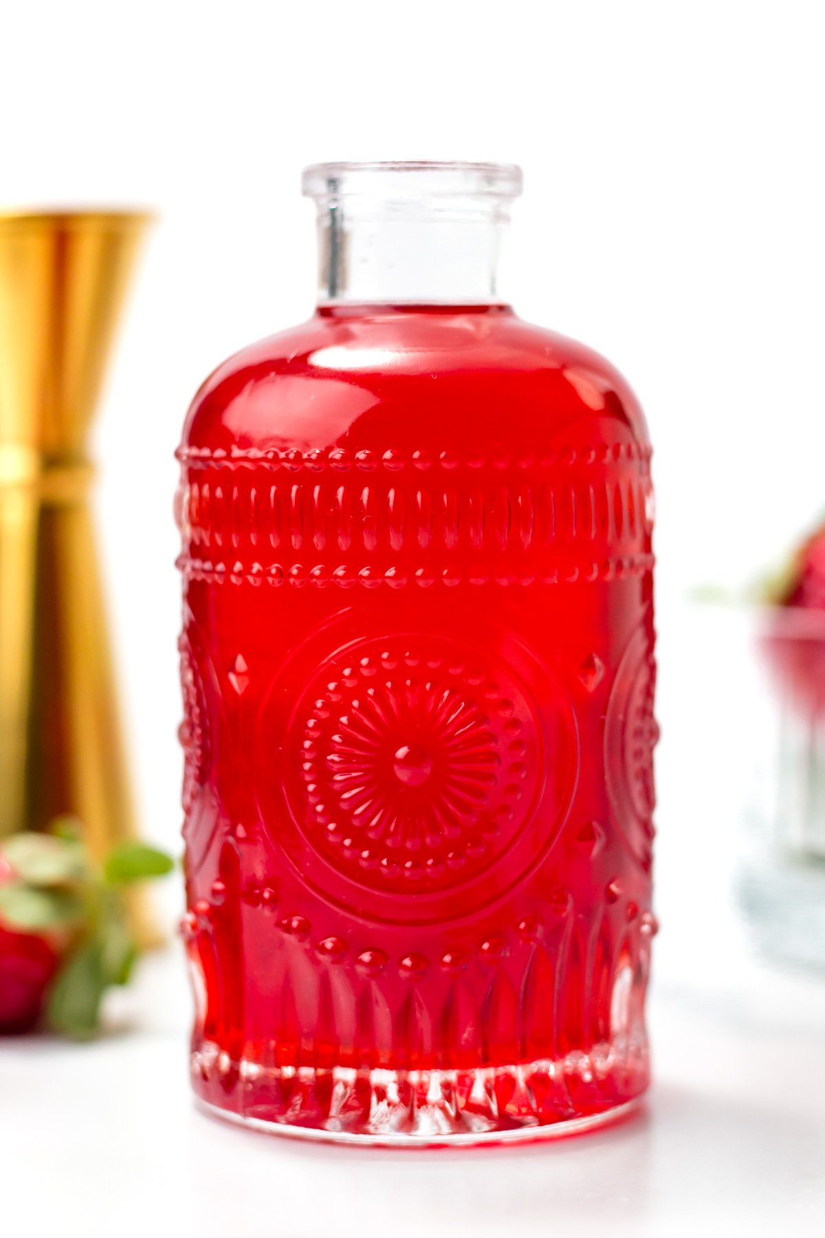 A clear glass bottle with an ornate pattern is filled with bright red liquid, set against a white background with a gold jigger and strawberries in the background.