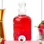 A decorative glass bottle filled with bright red strawberry syrup is surrounded by fresh strawberries and a gold jigger, with a label reading "Strawberry Syrup.
