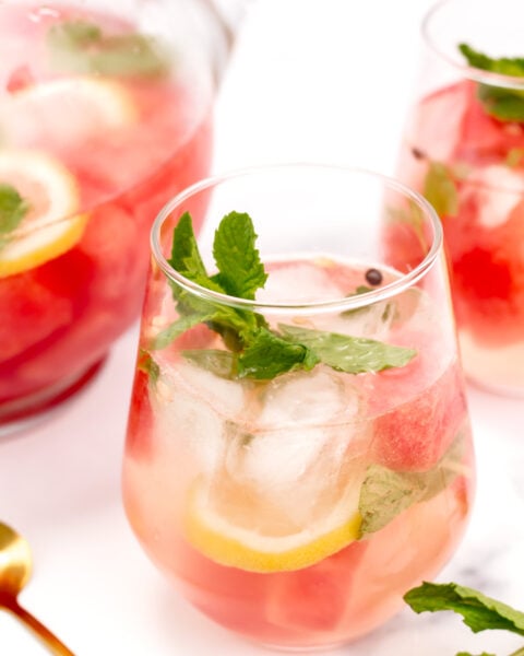 A glass of iced pink drink with lemon slices, mint leaves, and large ice cubes, next to a pitcher and another glass of the same beverage.