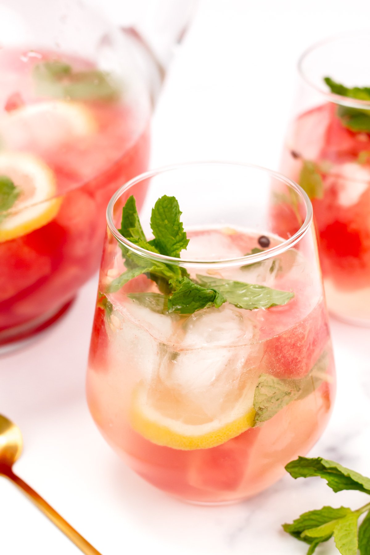 A glass of iced pink drink with lemon slices, mint leaves, and large ice cubes, next to a pitcher and another glass of the same beverage.