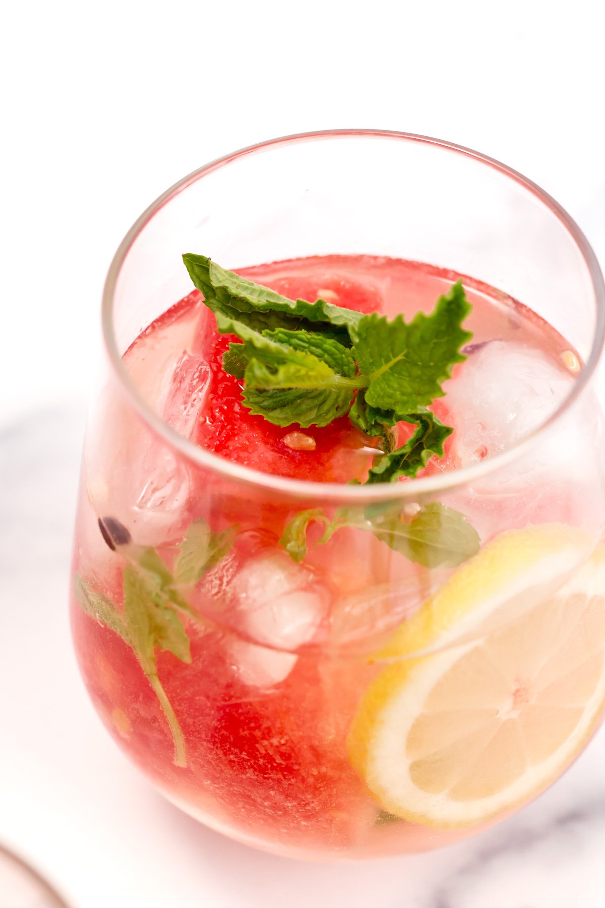 A glass of iced beverage with watermelon balls, a slice of lemon, fresh mint leaves, and clear ice cubes.
