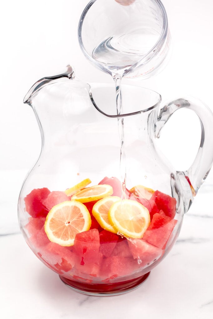 A glass pitcher filled with watermelon cubes and lemon slices, with water being poured in from a clear measuring cup.