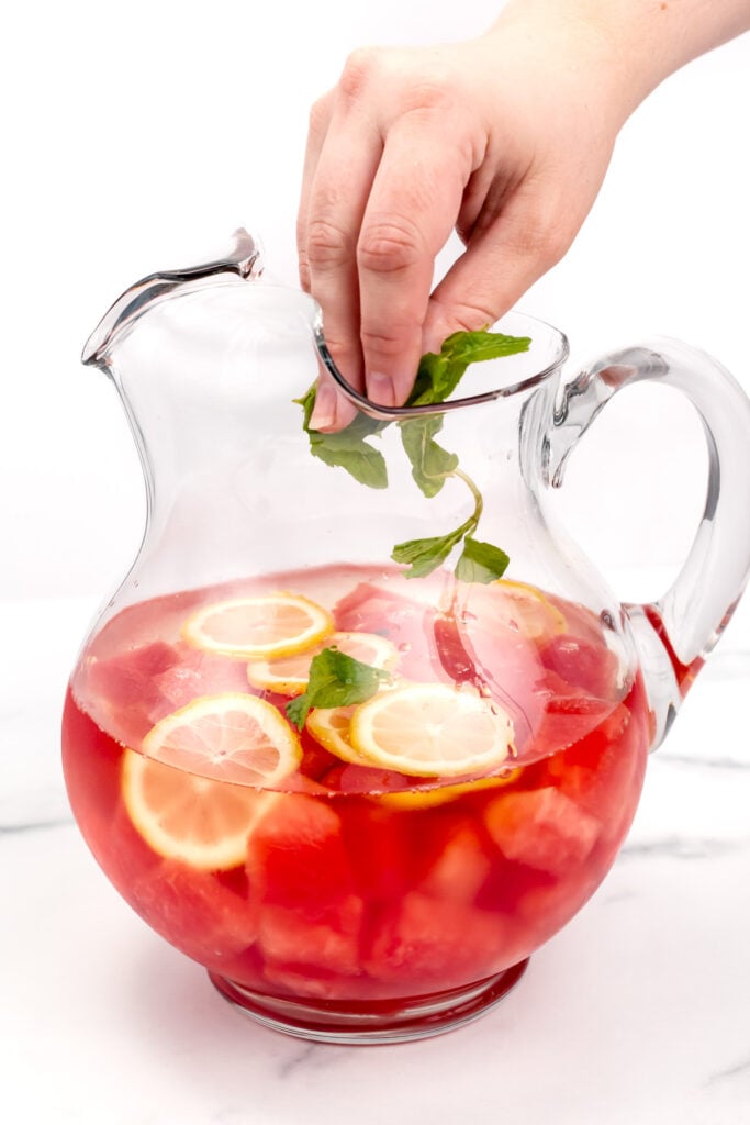 A hand adds fresh mint leaves to a glass pitcher filled with a red beverage, lemon slices, and watermelon chunks.