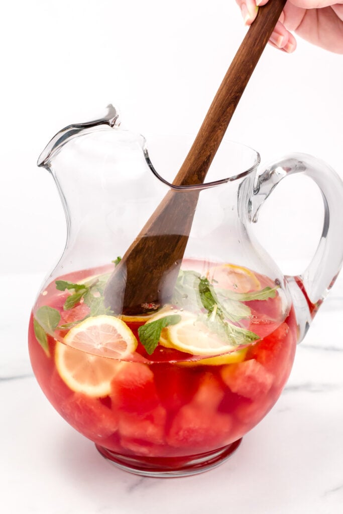 A glass pitcher filled with red fruit punch, lemon slices, mint leaves, and ice, being stirred with a wooden spoon.