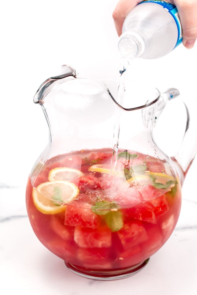 A hand pours sparkling water into a glass pitcher filled with watermelon cubes, lemon slices, mint leaves, and pink liquid.
