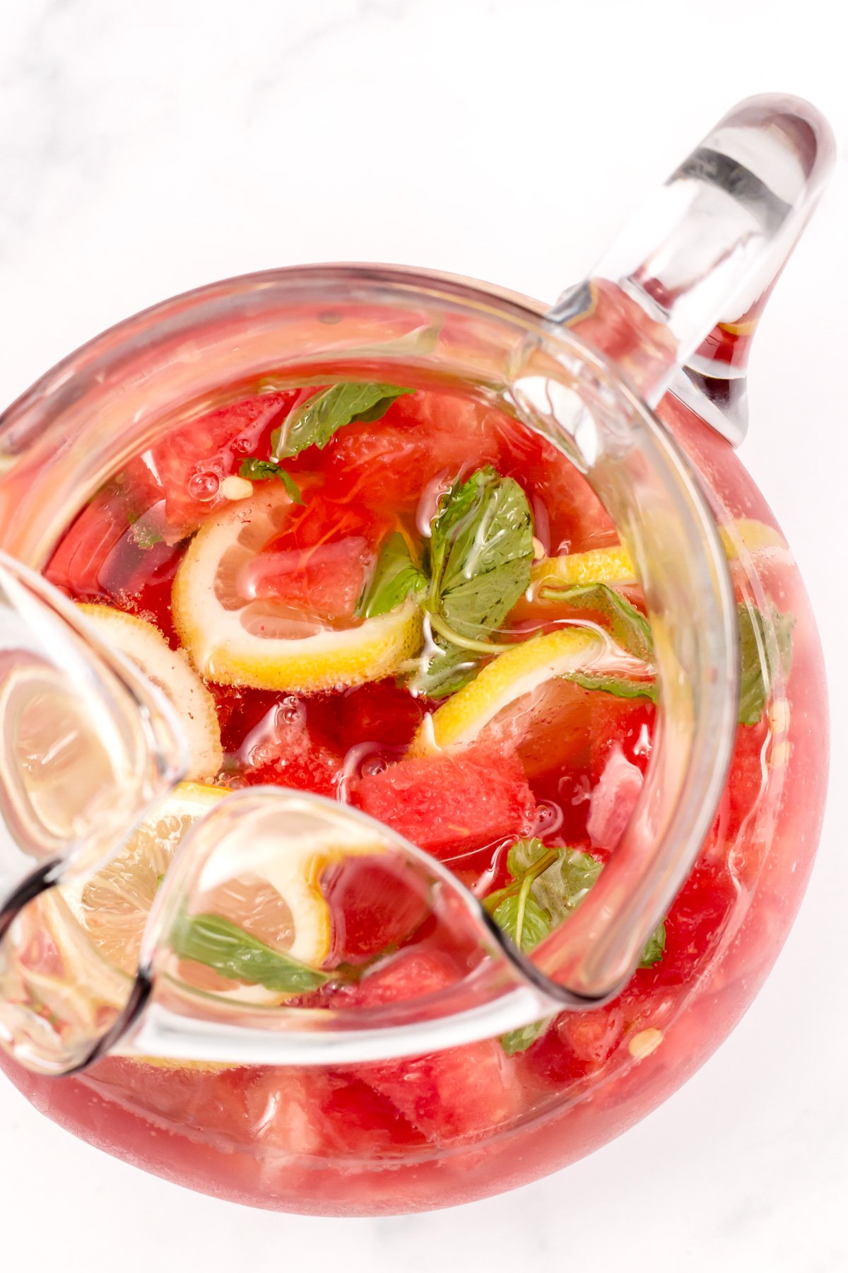 A glass pitcher filled with a pink beverage containing lemon slices, watermelon chunks, and fresh mint leaves on a white surface.