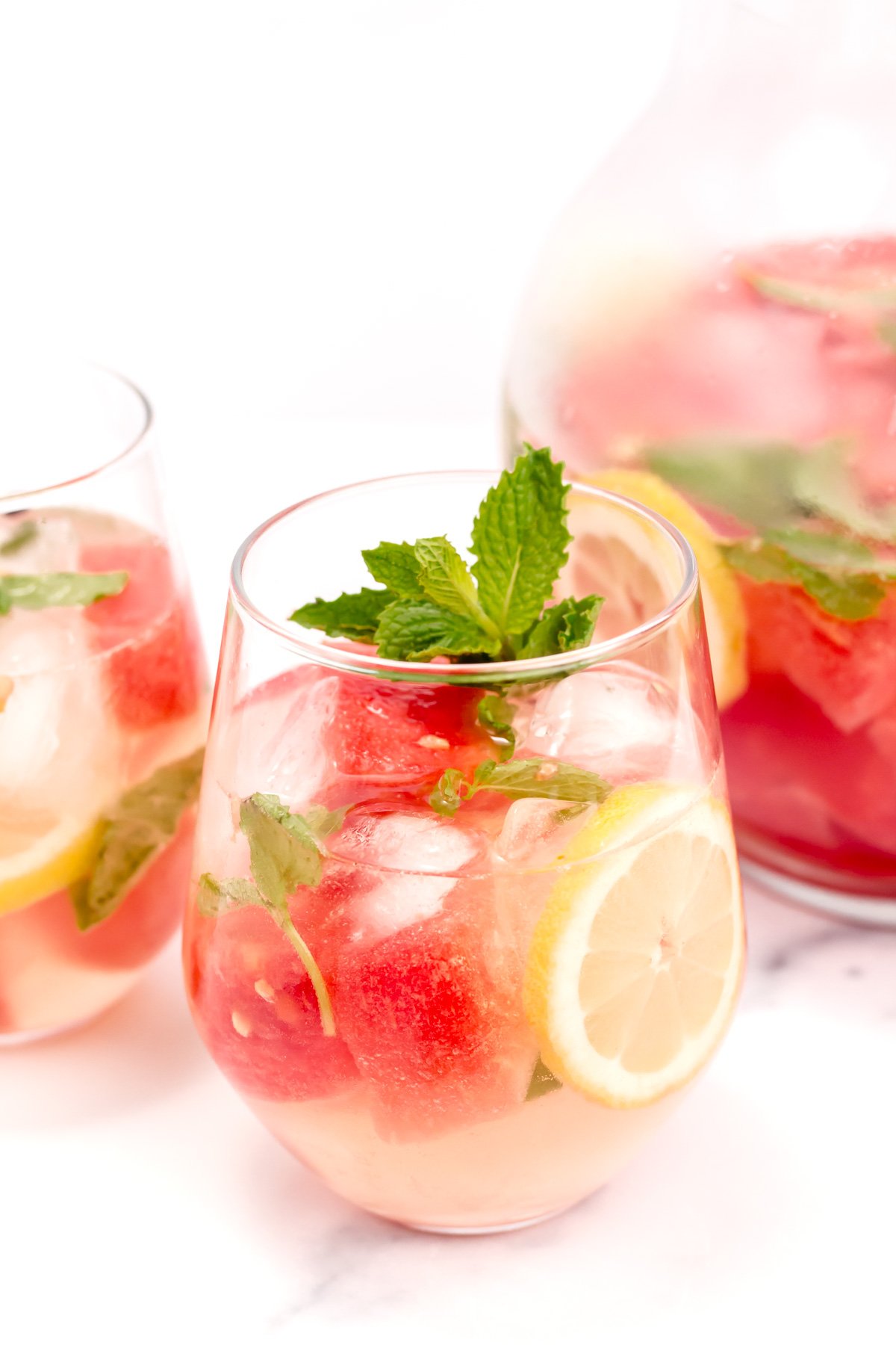 A glass of iced fruit-infused water with watermelon cubes, lemon slices, mint leaves, and ice, with a pitcher of the same drink in the background.