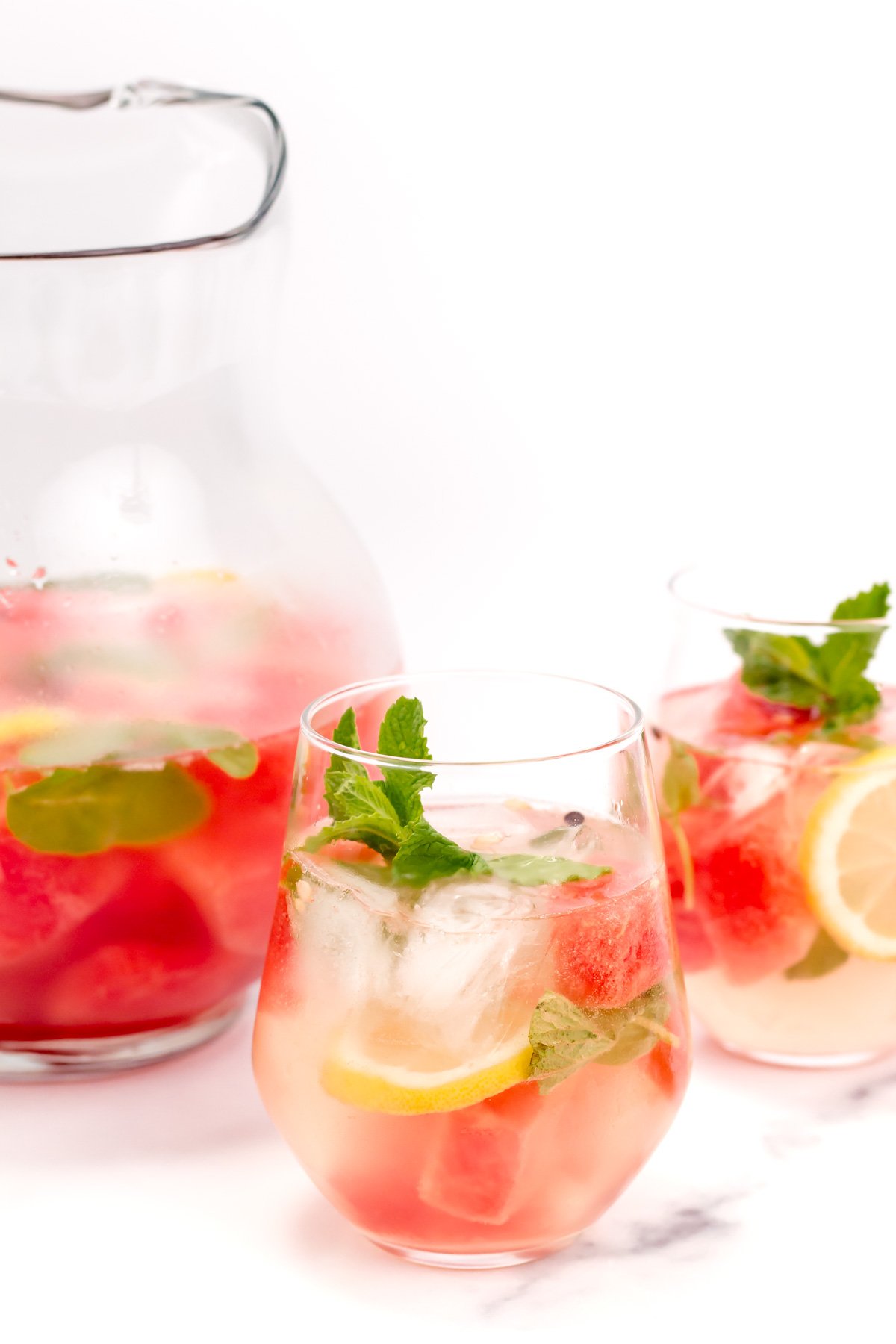 A glass pitcher and two glasses of fruit-infused water with lemon slices, watermelon cubes, ice, mint leaves, and clear liquid on a white background.