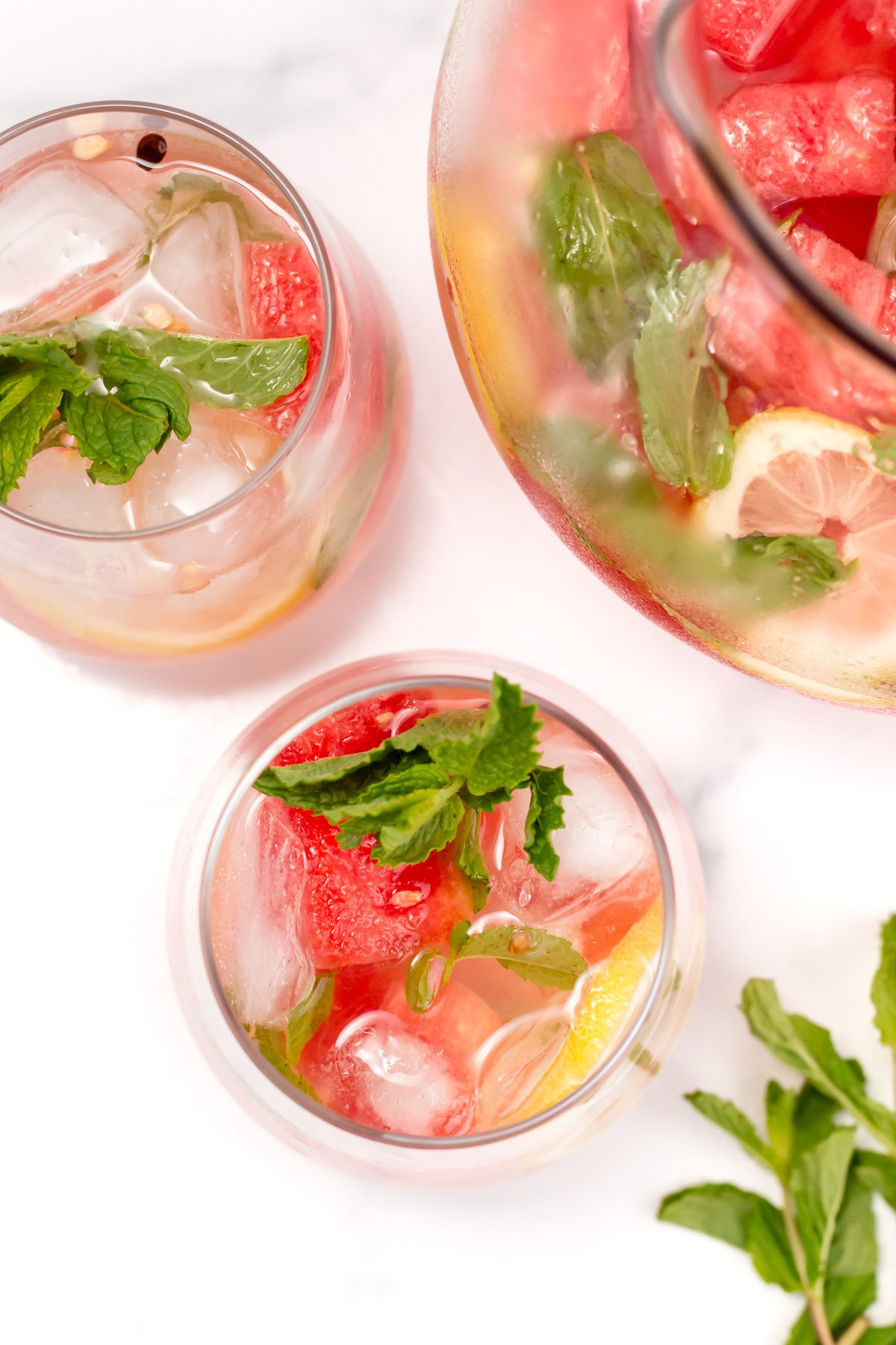 A top view of two glasses and a pitcher filled with a pink drink containing ice, watermelon chunks, lemon slices, and fresh mint leaves.