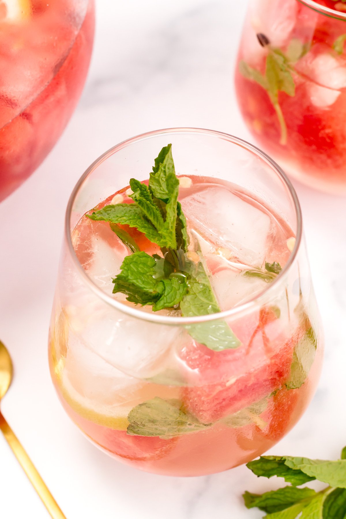 A glass of pink iced beverage with mint leaves and watermelon pieces, set on a white surface with part of another glass and a gold spoon visible.
