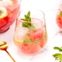 A glass of citrus watermelon sangria with ice, watermelon cubes, lemon slices, and mint leaves sits on a white surface next to a gold spoon.
