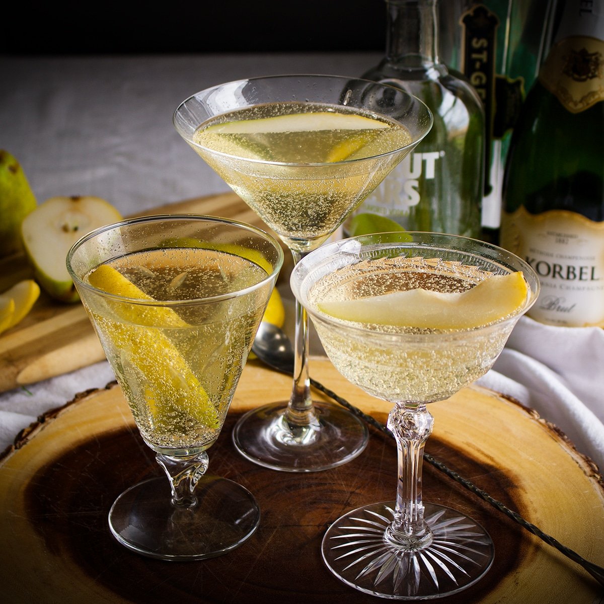 Three glasses of sparkling cocktails garnished with lemon peels sit on a wooden tray, surrounded by bottles of alcohol and sliced pears.