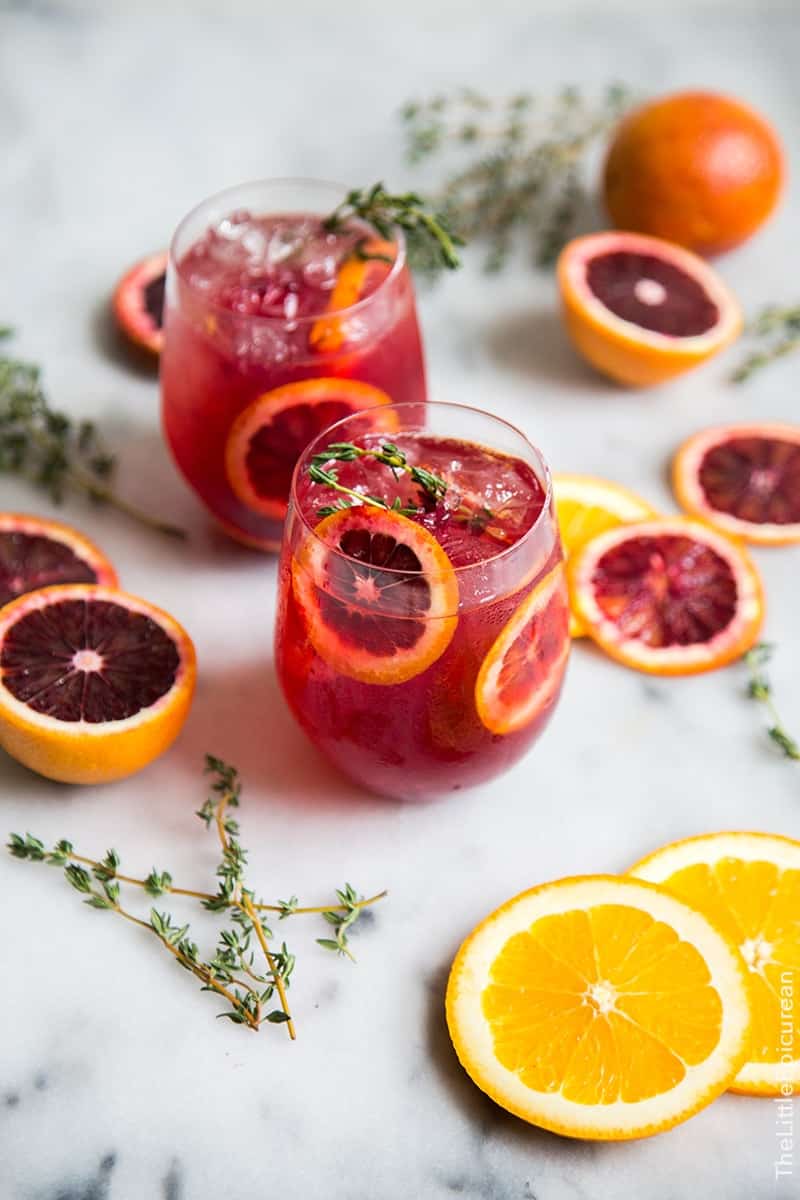 Two glasses of red citrus drink with ice, garnished with orange slices and thyme, surrounded by sliced oranges, blood oranges, and sprigs of thyme on a marble surface.
