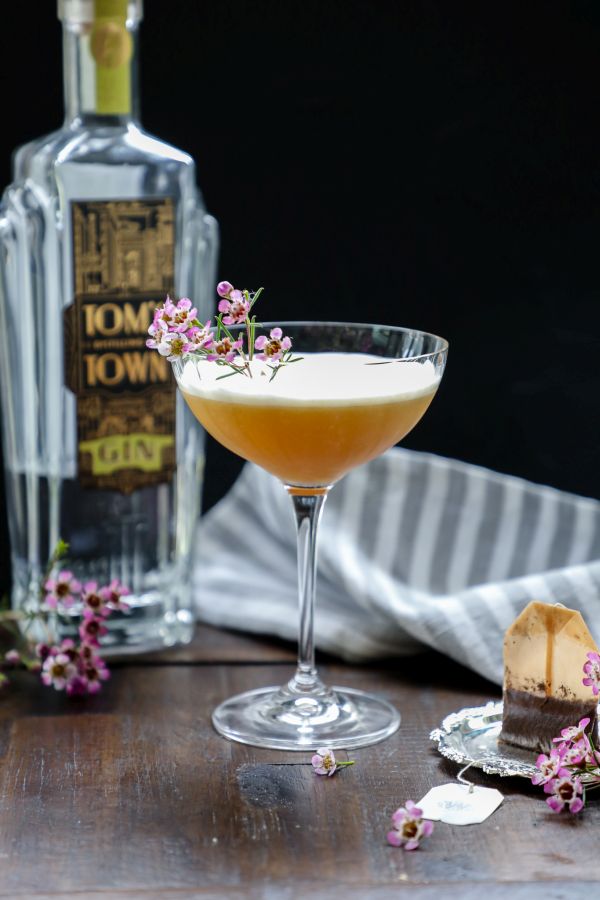 A cocktail garnished with small pink flowers sits in a coupe glass, next to a bottle of gin, a striped cloth, and a used tea bag on a saucer.