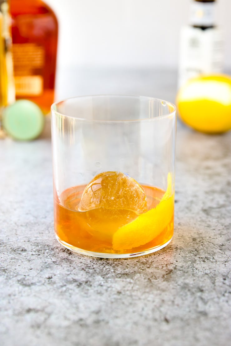 A glass of whiskey with a large round ice cube and a twist of lemon peel, placed on a gray surface. Bottles and a lemon are blurred in the background.
