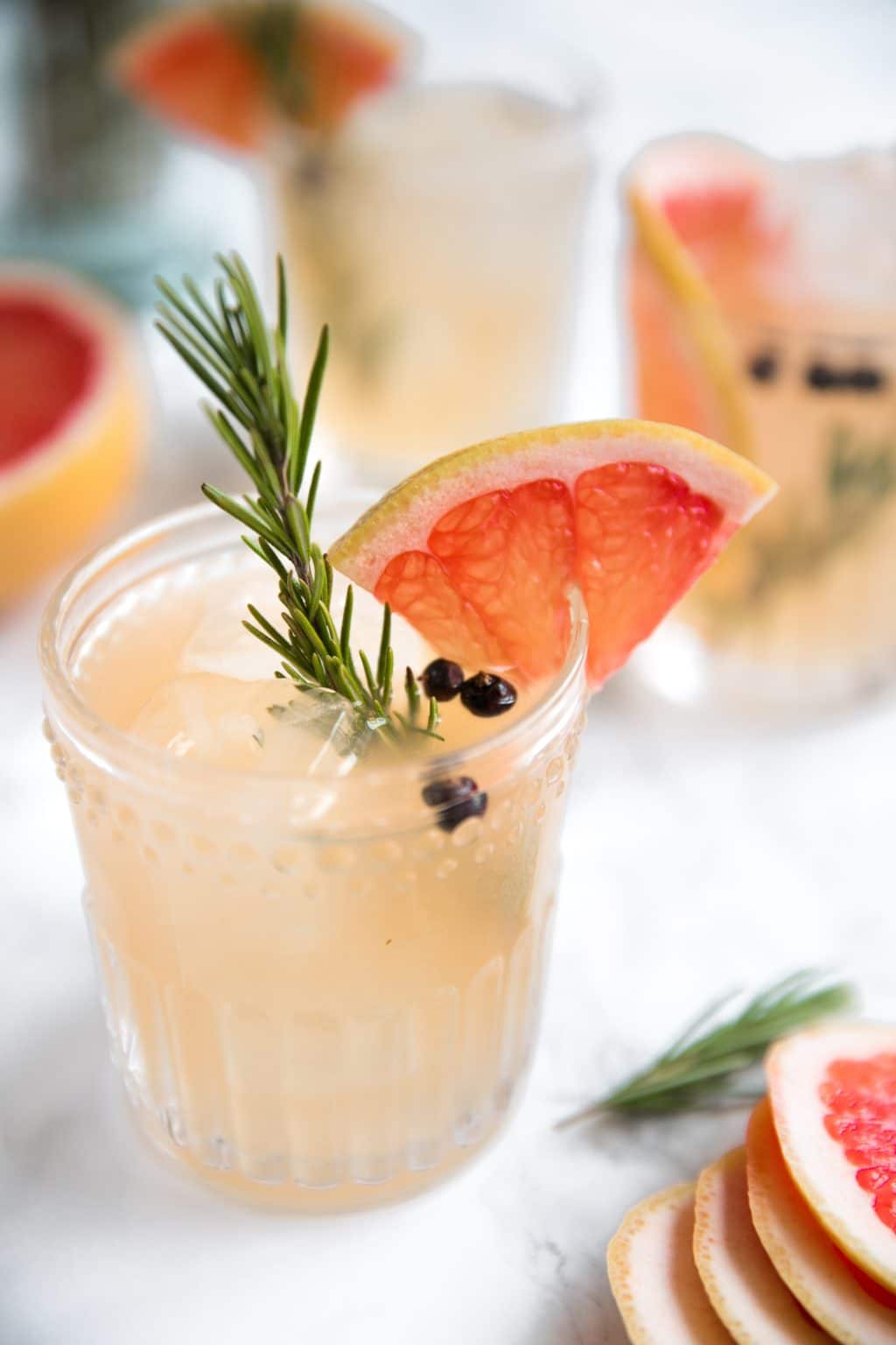 A glass of grapefruit cocktail garnished with a grapefruit slice, rosemary sprig, and juniper berries, with more drinks and grapefruit slices in the background.