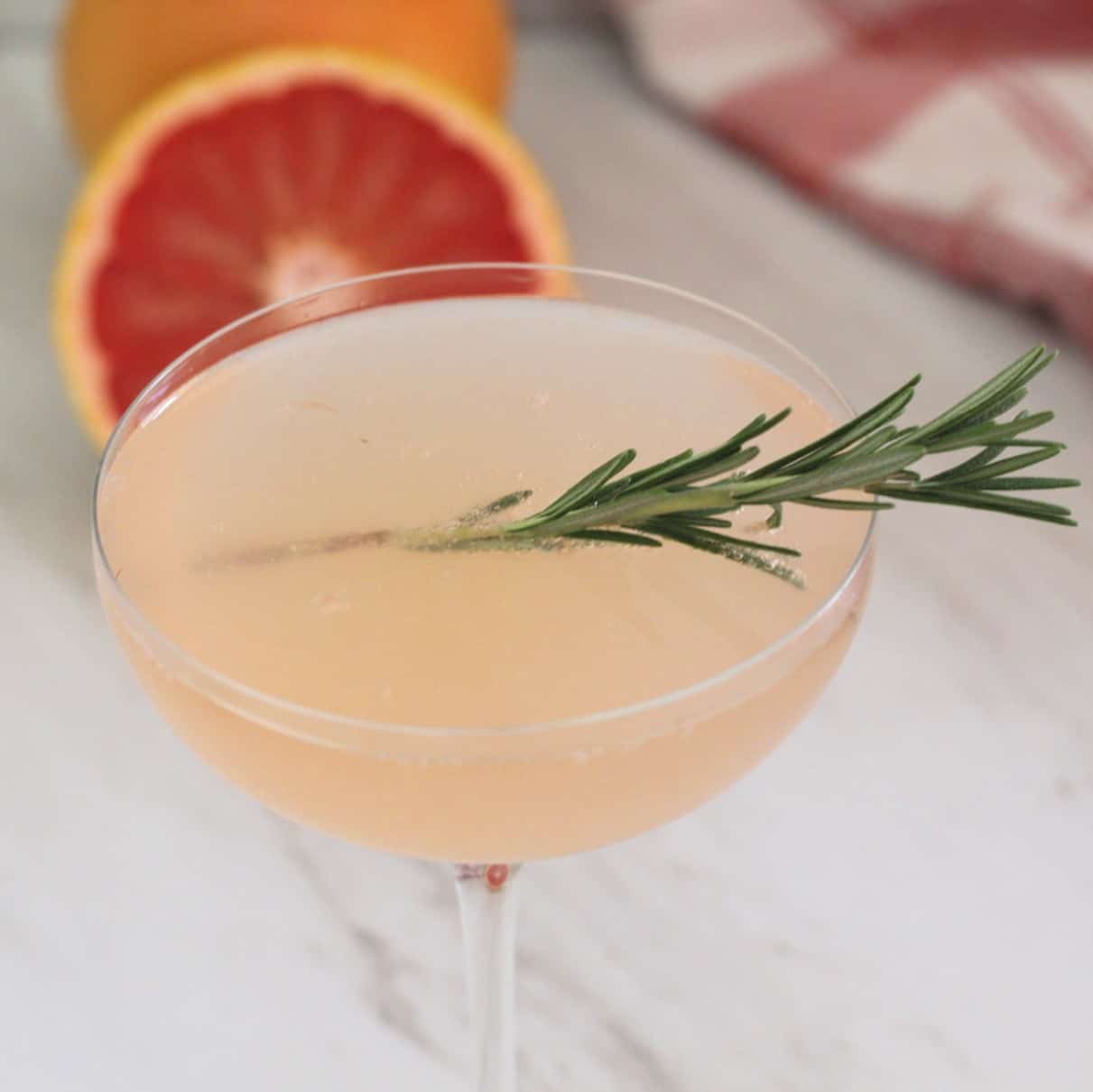 A pale pink cocktail garnished with a sprig of rosemary, with a halved grapefruit and a red and white cloth in the background.