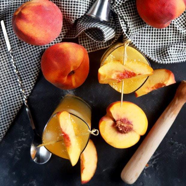 Overhead view of fresh peaches, two cocktails garnished with peach slices, a muddler, a spoon, and a checkered cloth on a dark surface.