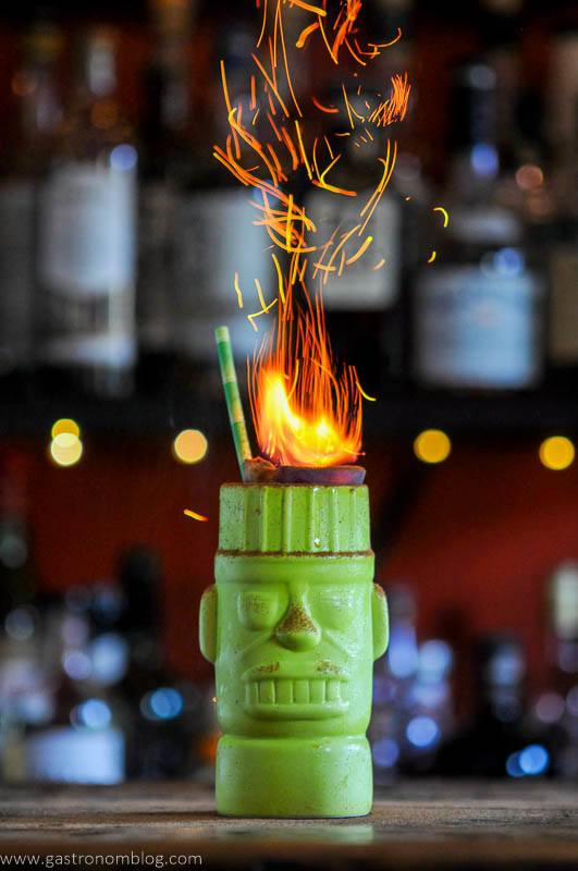A green tiki mug with a sculpted face holds a flaming garnish and a straw, with sparks rising above, set against a blurred background of liquor bottles.
