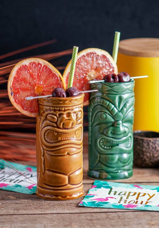 Two tiki mugs, one brown and one green, filled with cocktails, garnished with grapefruit slices and cherries, with straws and colorful napkins nearby.