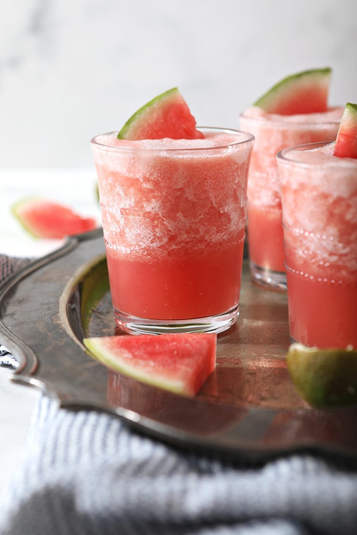 A glass of blended watermelon drink garnished with a watermelon wedge, placed on a metal tray with additional watermelon slices and other glasses in the background.