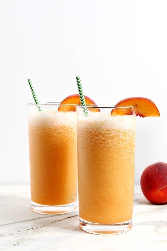 Two tall glasses of peach-colored slush with green striped straws, each garnished with a peach slice; a whole peach is on the side.
