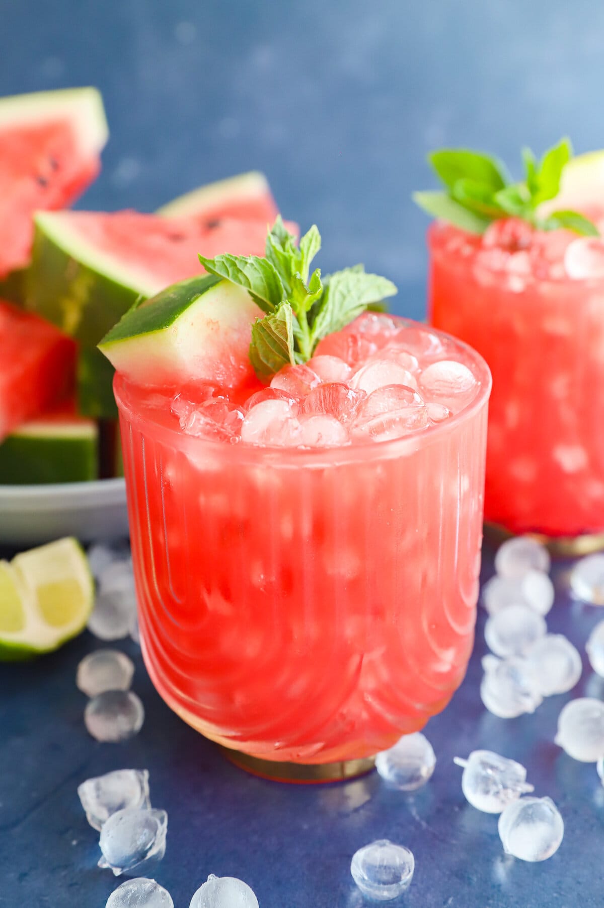 A glass of watermelon cocktail with ice, garnished with a watermelon slice and mint, sits on a blue surface with ice cubes and watermelon slices nearby.