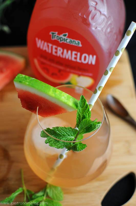 A glass of watermelon juice with a polka dot straw, garnished with a watermelon slice and fresh mint; a bottle of Tropicana Watermelon sits in the background.
