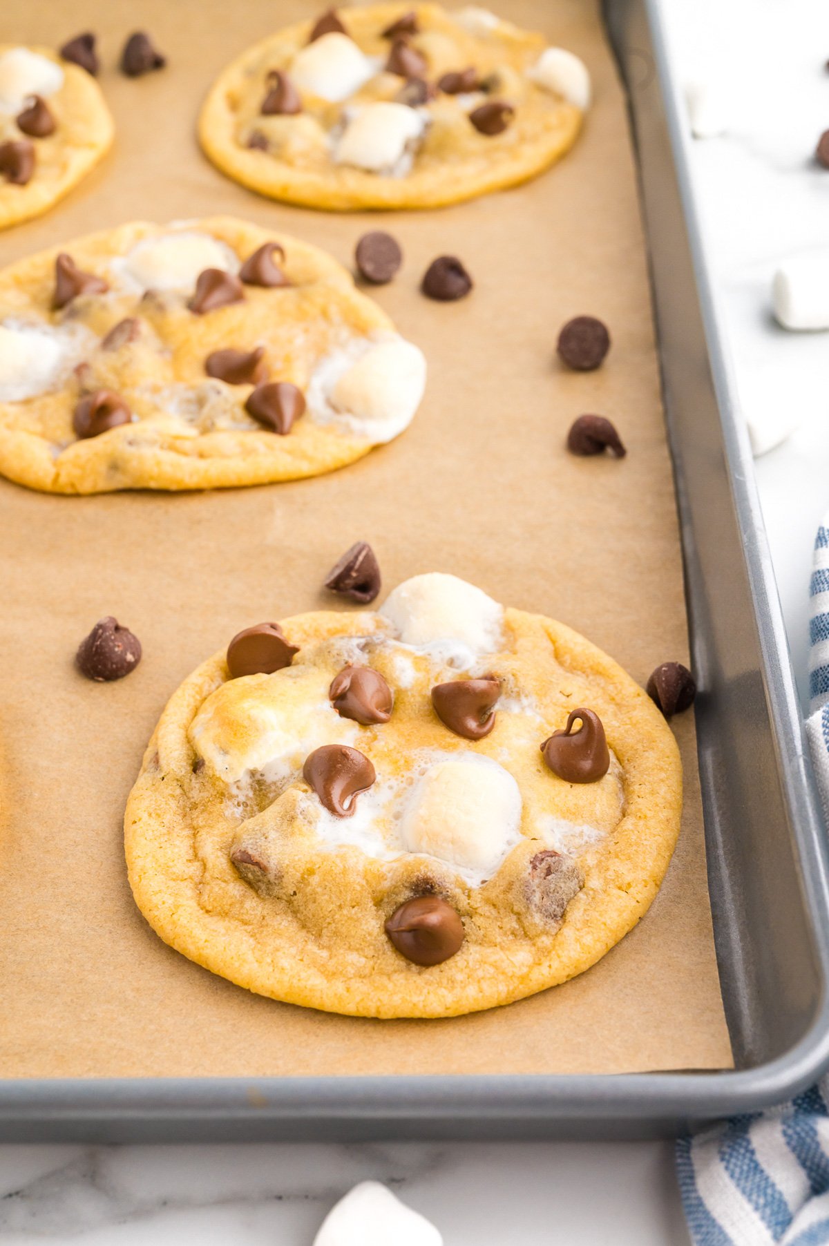 Chocolate chip and marshmallow cookies on a parchment-lined baking sheet, with a few chocolate chips and marshmallows scattered nearby.
