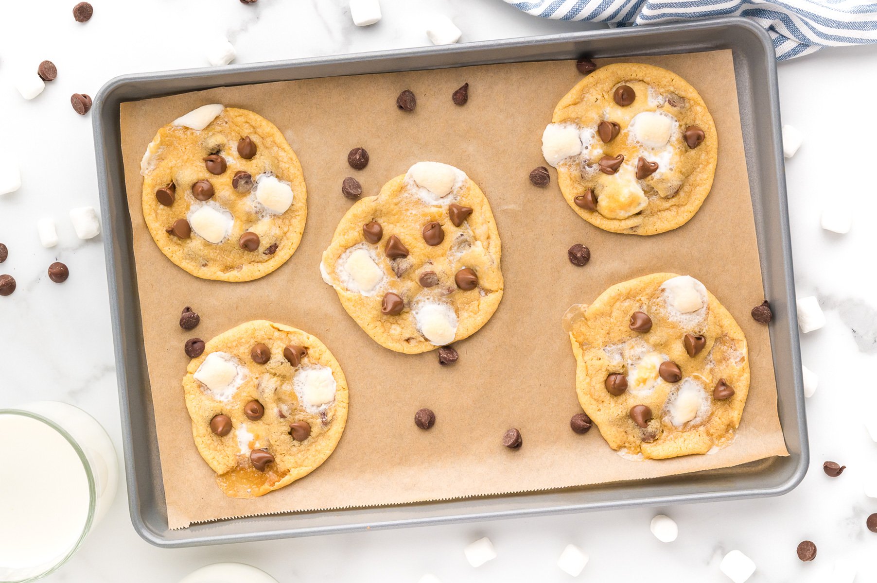 A baking tray with five cookies topped with marshmallows and chocolate chips, placed on parchment paper, surrounded by loose chocolate chips and mini marshmallows.