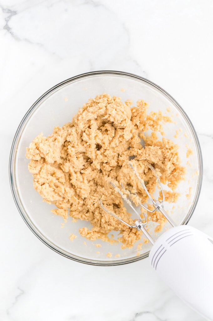 A glass bowl containing cookie dough being mixed with an electric hand mixer on a white marble surface.