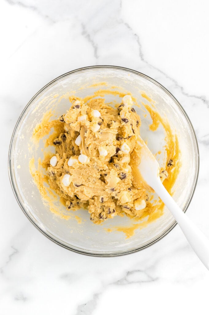 A glass bowl filled with cookie dough containing chocolate chips and white chocolate chips, with a white spatula resting inside.
