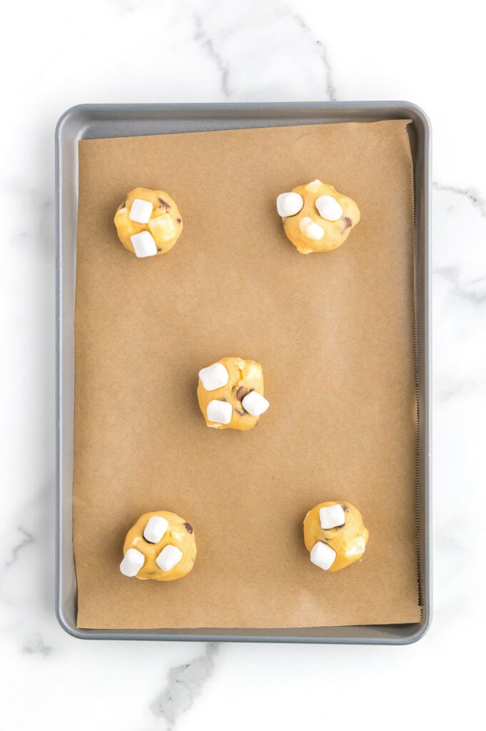 A baking sheet lined with parchment paper holds five evenly spaced balls of cookie dough topped with mini marshmallows, ready to be baked.